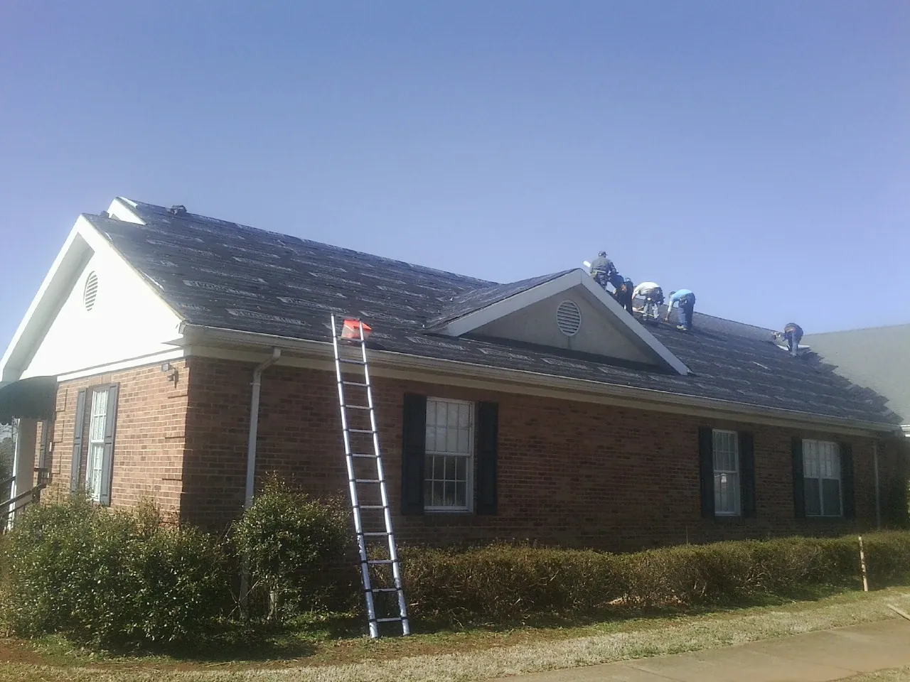 Crew tearing shingles off a brick ranch roof with a ladder set — Metal Roof Repair work by our crew in Mc Bee