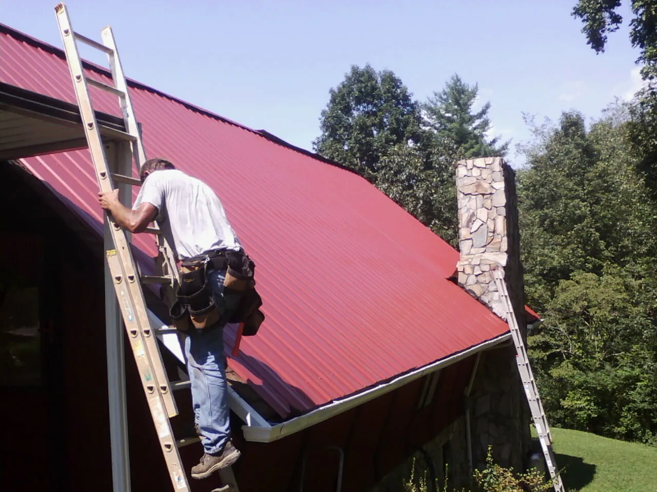 Roofer on a ladder beside a red metal roof and stone chimney — Soffit & Fascia Repair by Roofing Brothers in Mc Bee